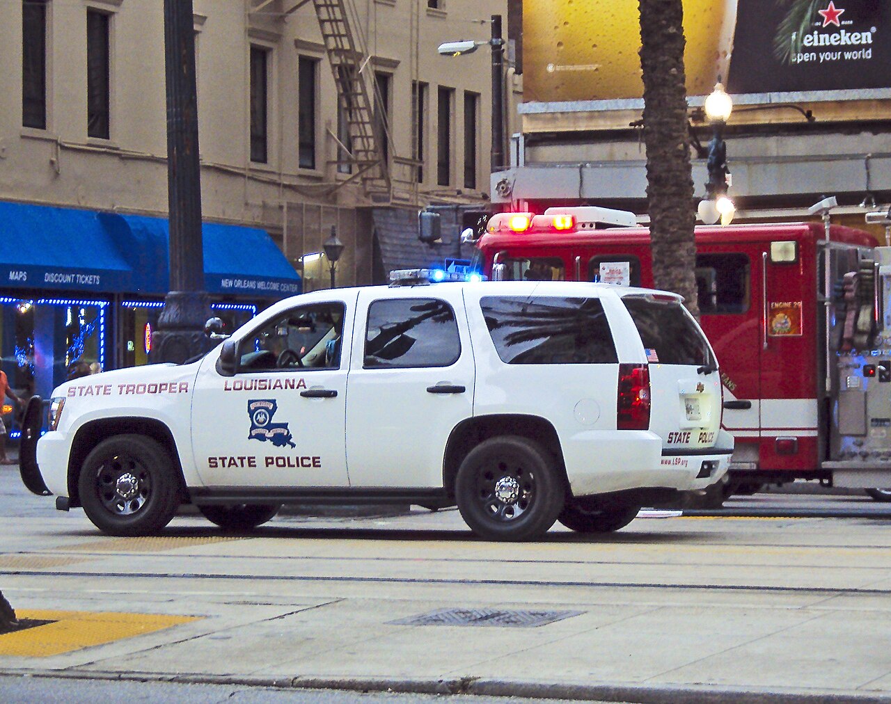 A white Louisiana State Police SUV marked State Trooper parked on a city street, a fire truck behind it.