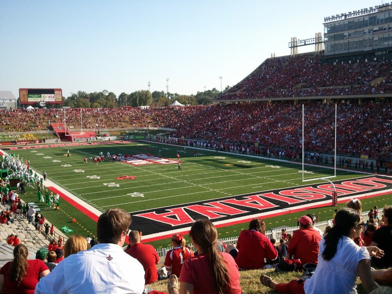 Fans in red sit on a grassy hill above a packed football stadium, the end zone painted LOUISIANA.