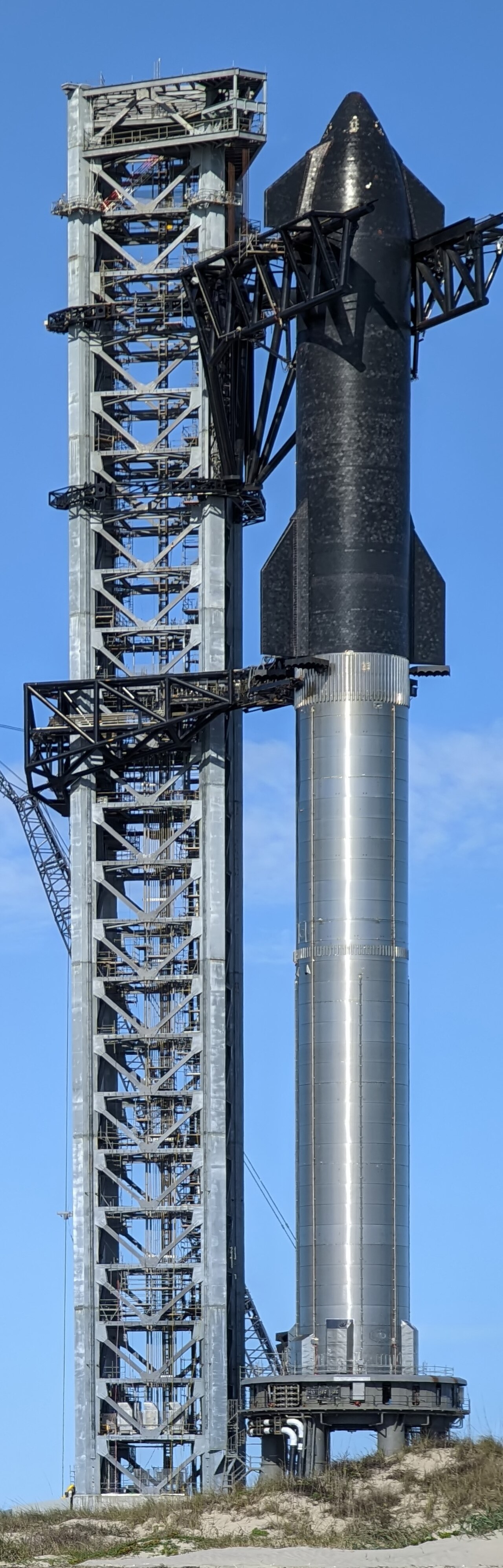 A stainless-steel SpaceX Starship rocket stacked on its booster beside the launch tower, under a blue sky.
